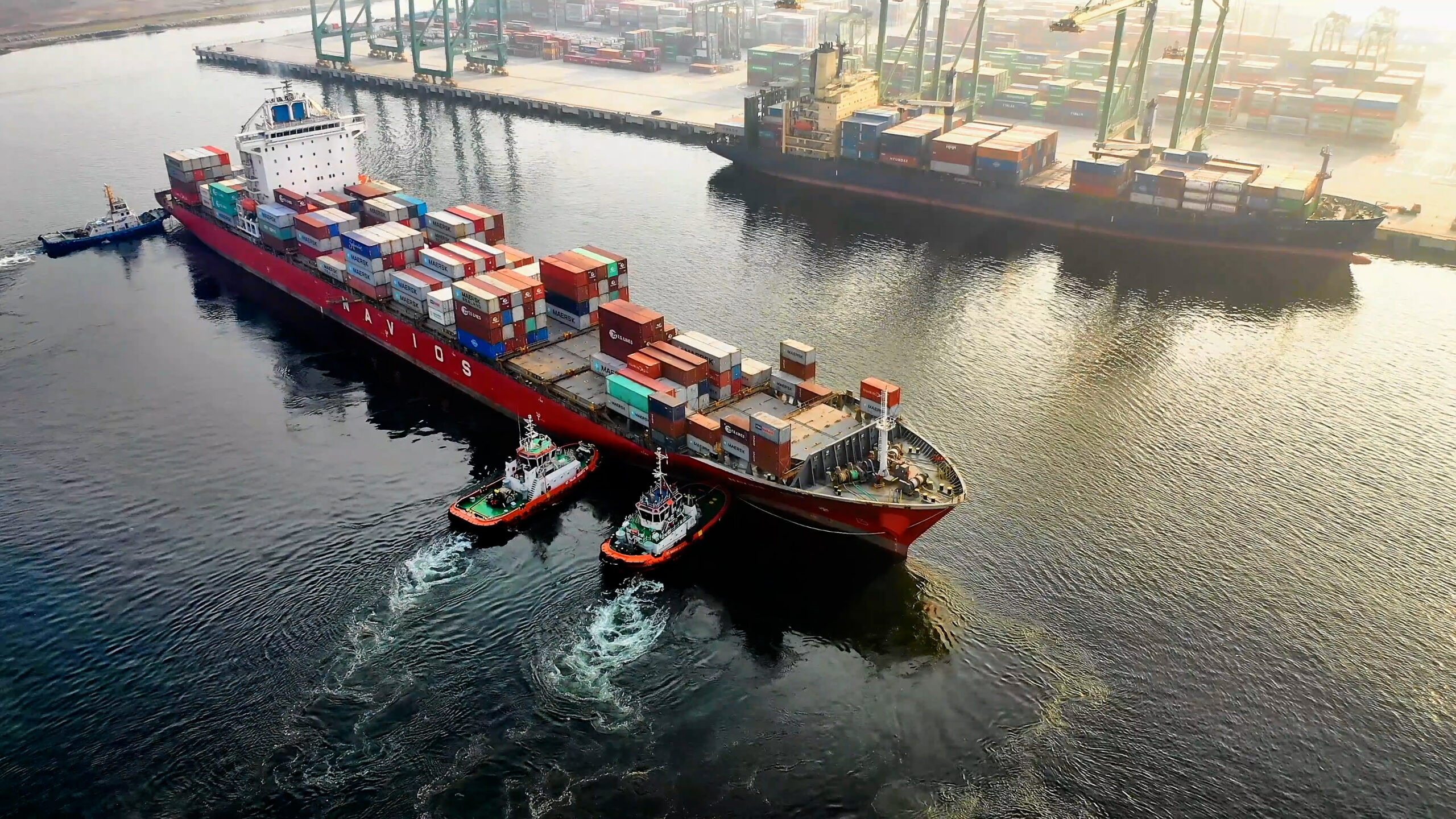 Aerial view of a container ship floating on the water. The ship seems to be in the middle of a voyage, and it is likely transporting large quantities of goods in shipping containers. The image captures the vast size and scope of the ship, as well as the surrounding water and landscape.